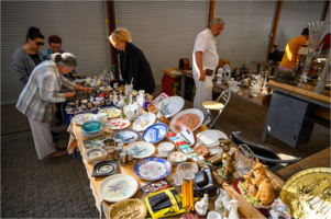 Na zdjęciu stół z rzeczami, znajdują się na nim talerze oraz inne bibeloty, obok ludzie przyglądający się towarowi.